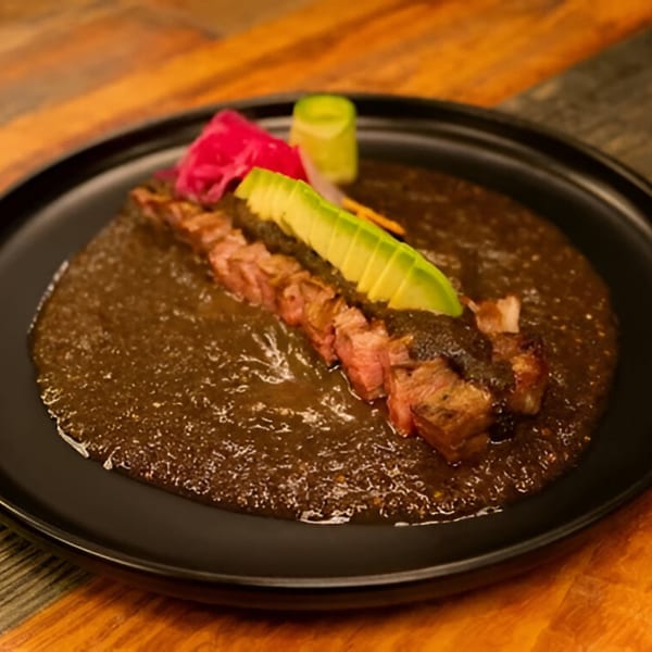Mexican dish with sliced meat, avocado, and pickled onions in a dark sauce on a black plate.